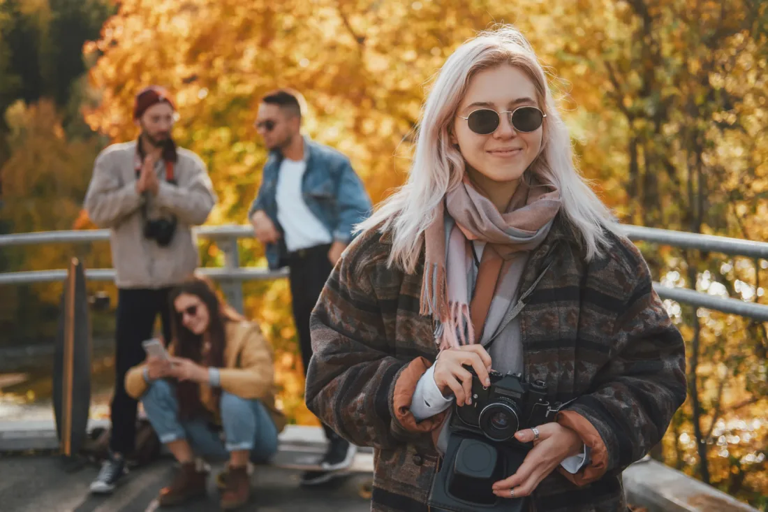 Fată blondă îmbrăcată la modă, cu ochelari de soare și aparat de fotografiat, se distrează cu prietenii ei în parcul public de toamnă.