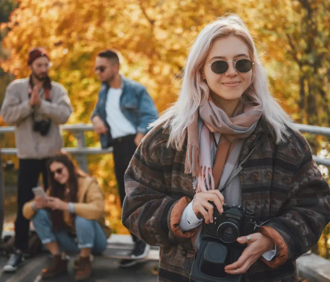 Fată blondă îmbrăcată la modă, cu ochelari de soare și aparat de fotografiat, se distrează cu prietenii ei în parcul public de toamnă.